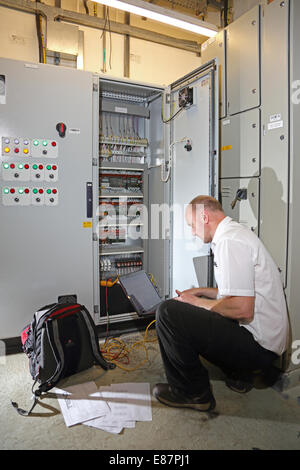A building services engineer monitors the electrical control cabinet in the plant room of a large office block using a computer Stock Photo