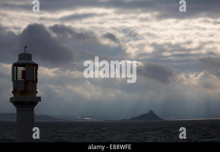 sun rays beaming down on Michaels Mount, Cornwall through stormy skies with harbour lighthouse Penzance in the foreground April Stock Photo