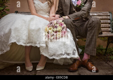 A bride & groom wearing a vintage style tweed suit& 50's style wedding,sitting on a bench, holding hands & a bouquet of roses Stock Photo