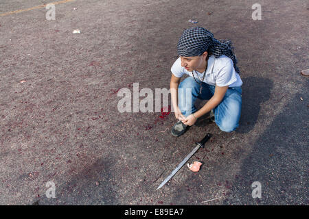 A young Shiite Muslim boy with a sword, ties his shoelaces on a bloody street, during the Day of Ashura in Nabatieh, Lebanon. Stock Photo