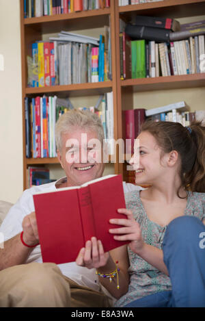 Grandfather and granddaughter reading book on couch in living room, smiling Stock Photo