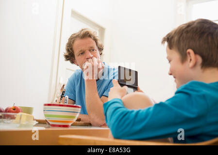 Father looking at son playing video game at breakfast table Stock Photo