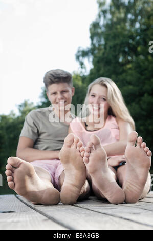 Smiling teenage couple sitting on a jetty at lake Stock Photo