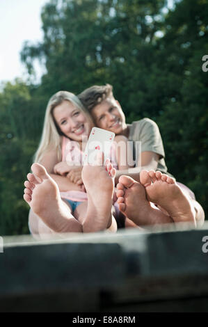 Teenage couple sitting on a jetty at lake, girl holding an ace between her toes Stock Photo