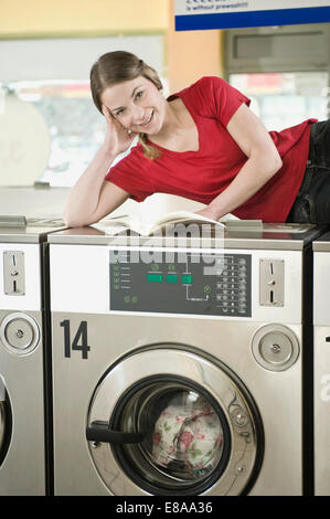 Portrait of young woman reading book on top of machine, smiling Stock Photo