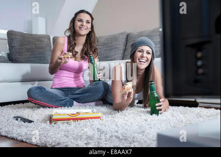 Two female friends side by side on carpet at home watching television
