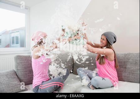 Two female friends sitting on couch at home having pillow fight Stock Photo