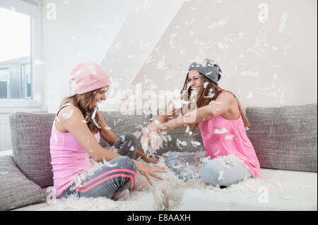 Two female friends sitting on couch at home having pillow fight Stock Photo