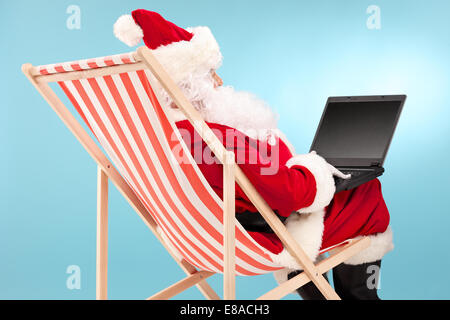 Santa Claus working on a laptop seated in a sun lounger on a blue background, rear view Stock Photo