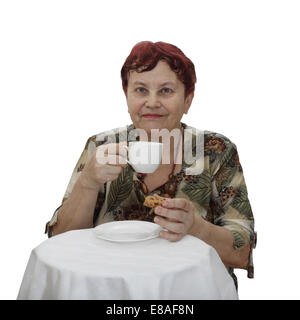 Elderly woman sits at table with cup of tea and cookie in hands isolated on white background Stock Photo