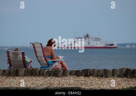 A couple relaxes on Calshot beach looking out to the Solent as a red Funnel boat goes to the isle of Wight Stock Photo