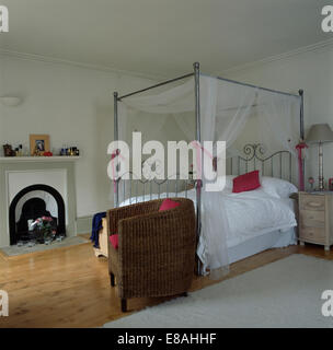 Wicker tub chair beside chrome four-poster bed with white voile drapes and white duvet in white country bedroom Stock Photo