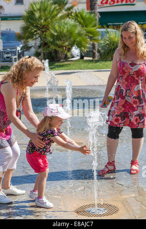 Young girl playing in water spout fountain with mother. Stock Photo