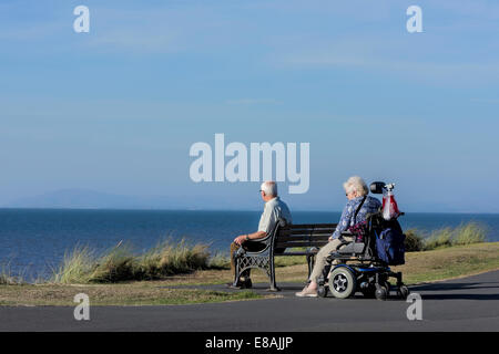 Man sitting on bench with wife in wheelchair on a coastal path in Blackpool, Lancashire, UK Stock Photo