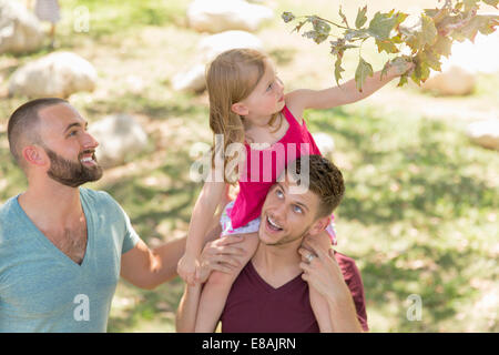 Young girl sitting on fathers shoulders reaching for tree leaf in park Stock Photo