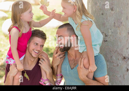 Two sisters sitting on fathers shoulders in park Stock Photo