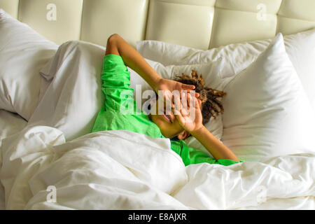 Sleepy boy lying in bed shielding face with hands Stock Photo