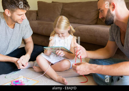 Male couple and daughter threading picture books on sitting room floor Stock Photo