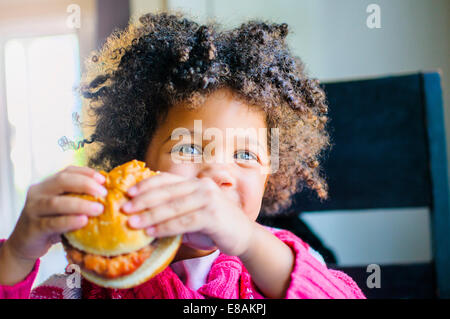 Portrait of cute girl eating hamburger in kitchen Stock Photo