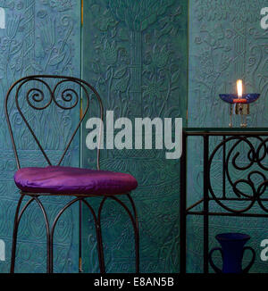 Purple silk cushion on wrought iron chair in front of textured turquoise folding screen Stock Photo
