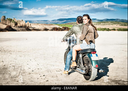 Rear view of young couple riding motorcycle on arid plain, Cagliari, Sardinia, Italy Stock Photo