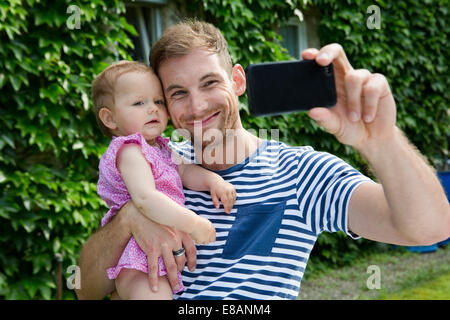 Mid adult man with baby daughter taking selfie on smartphone in garden Stock Photo