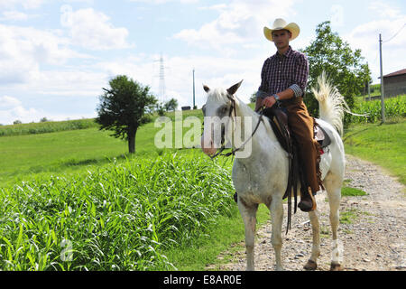 Portrait of young man in cowboy gear riding horse on rural road Stock Photo