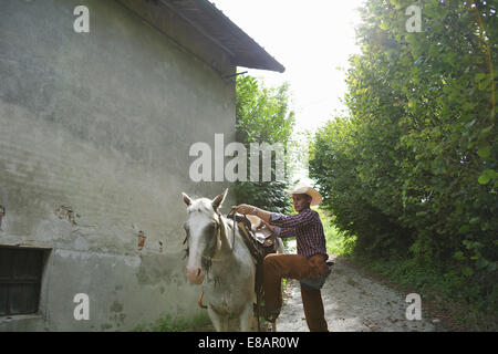 Young man in cowboy gear mounting horse outside barn