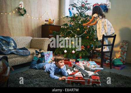 Girl arranging christmas tree whilst brother plays with xmas gifts on sitting room floor