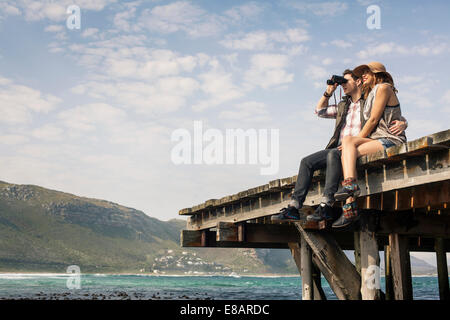Young couple sitting on edge of old pier looking through binoculars, Cape Town, Western Cape, South Africa Stock Photo