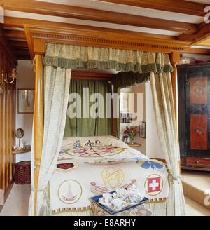 Patterned drapes on four-poster bed with antique patchwork quilt in traditional country bedroom with breakfast tray on stool Stock Photo