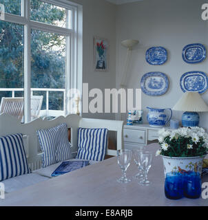 Collection of blue+white plates on wall of dining room with blue striped cushions on white settle in front of window