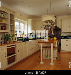 Stools beneath breakfast bar on small island unit in country kitchen with wooden flooring and cream painted units Stock Photo