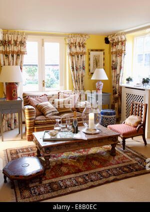 Brown striped sofa and floral curtains in yellow living room with Indonesian coffee table on patterned rug Stock Photo