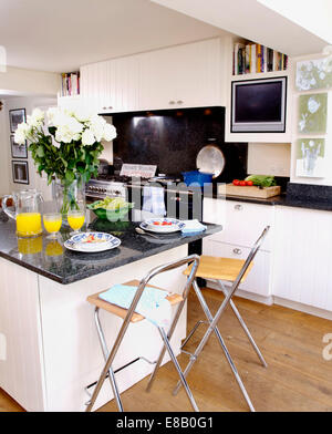 Stools at breakfast bar with vase of white roses and jug of orange juice on granite worktop in white country kitchen Stock Photo