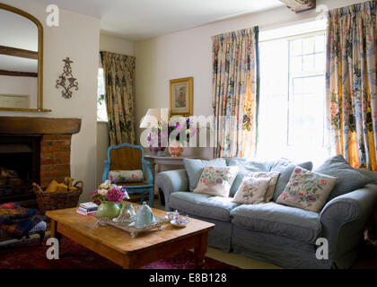 Blue sofa in front of window with floral curtains in country living room with tea set on wooden table Stock Photo