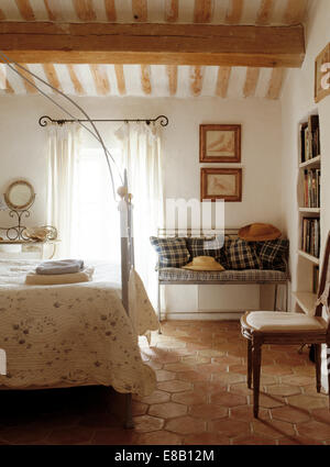 Simple wrought iron four poster bed in French country bedroom with checked cushions on settle and hexagonal tiled floor Stock Photo