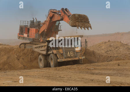 An orange Hitatchi excavator loads waste rock and soil into a Caterpillar mining truck in a large open cast African copper mine. Stock Photo