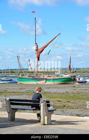 Woman relaxing beside the Blackwater estuary with Thames Sailing Barge 'Kitty' resting on mud flats at low tide beyond Stock Photo