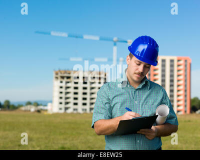 caucasian engineer standing, writing on clipboard, with buildings in construction on background Stock Photo
