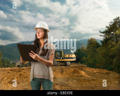 portrait of young caucasian female architect standing on construction site, holding clipboard and writing Stock Photo