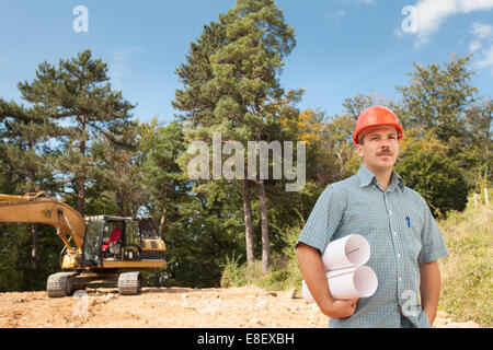 side view of standing engineer holding blueprints on construction site outdoors, with excavator in background Stock Photo