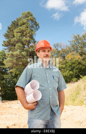 side view of standing engineer holding blueprints on construction site outdoors Stock Photo