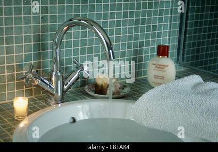 Close-up of lighted candle beside chrome tap pouring water into white basin in mosaic tiled bathroom Stock Photo