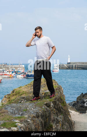 Young man standing on a tall rock, apparently to get a signal, and making a call from his cell mobile phone. Mevagissey. UK Stock Photo