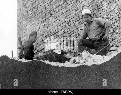 A building worker (right), under the surveillance of a border guard of the GDR (left), has a break during the demolition of a bordering house near Bernauer Street in Berlin, 2 June 1965. From 13 August 1961, the day of the building of the Berlin Wall,  to 9 November 1989, the day of the fall of the Berlin Wall, the Federal Republic of Germany and the GDR were separated by the iron curtain between West and East. Stock Photo