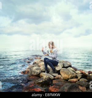 Woman sitting on rocks by ocean releasing a butterfly from a glass jar, Norway Stock Photo