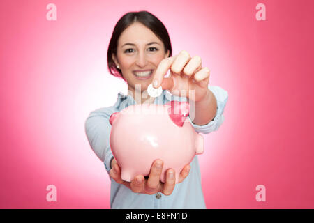 Portrait of a smiling young woman inserting a coin into a piggy bank Stock Photo