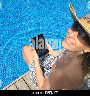 Smiling woman with digital tablet sitting at the edge of swimming pool Stock Photo
