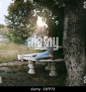 Woman sitting under a tree reading book with a steaming cup of tea on table, Spain Stock Photo